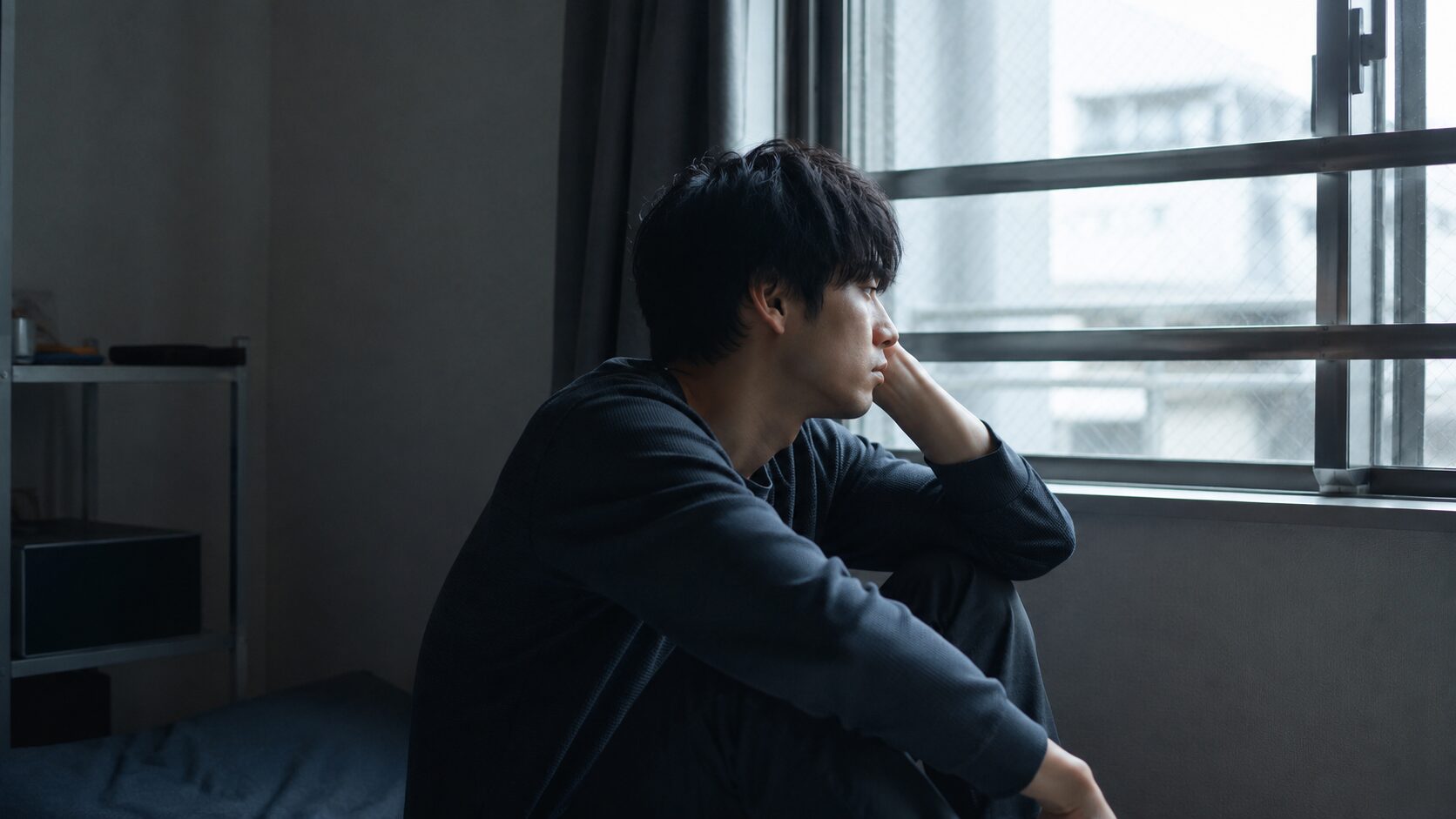 うつ病で働けないことに悩み、一人で窓の外を見つめる男性。「一生働けないかもしれない」という不安や将来への絶望感をイメージした写真。